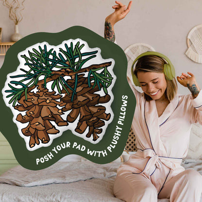 This image shows a photo of the pinecones-shaped pillow in a green organic shape. Inside the shape are the words "Posh your pad with plushy pillows". In the background, there is a photo of a woman in pajamas wearing headphones and sitting on a bed.