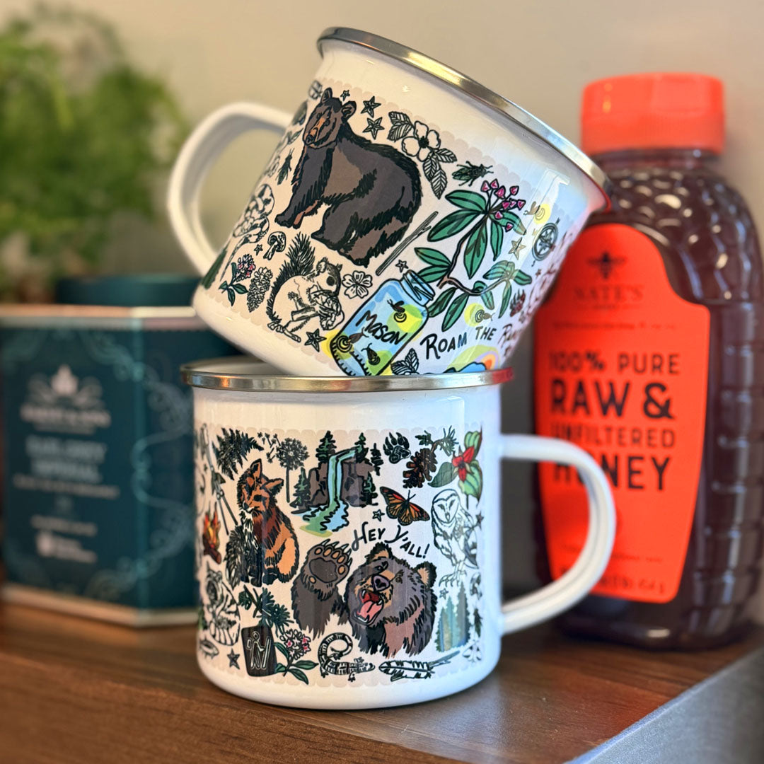 Two Mountain Hikes enamel mugs with bear and nature illustrations on a wooden kitchen shelf with a bottle of honey in the background.