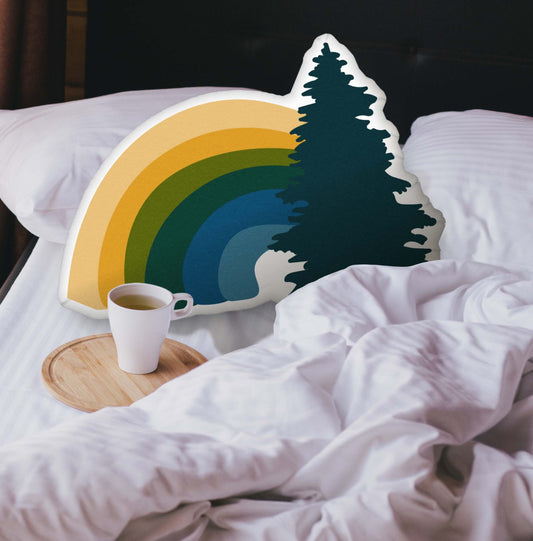 Rainbow-shaped pillow nestled in an unmade bed. There is a cup of coffee sitting on a wooden tray.