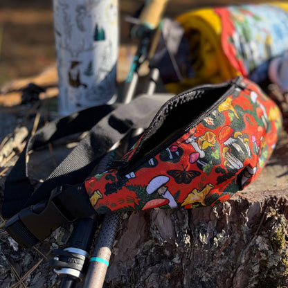 The red mushroom sling bag laying unzipped on a tree stump. Trekking poles, water bottle, and a blanket are in the background.