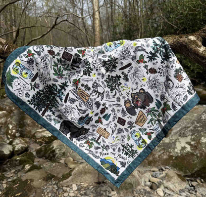 The Smoky Mountains bandana scarf is lying across a tree branch in the Smoky Mountains. You can see the detail of the illustrations on the bandana. The illustrations are iconic images which include a mason jar with fireflies, black bears, hiking symbols and trail markers, mountain laurel, wildflowers, foxes, waterfalls, pine trees and other plants.