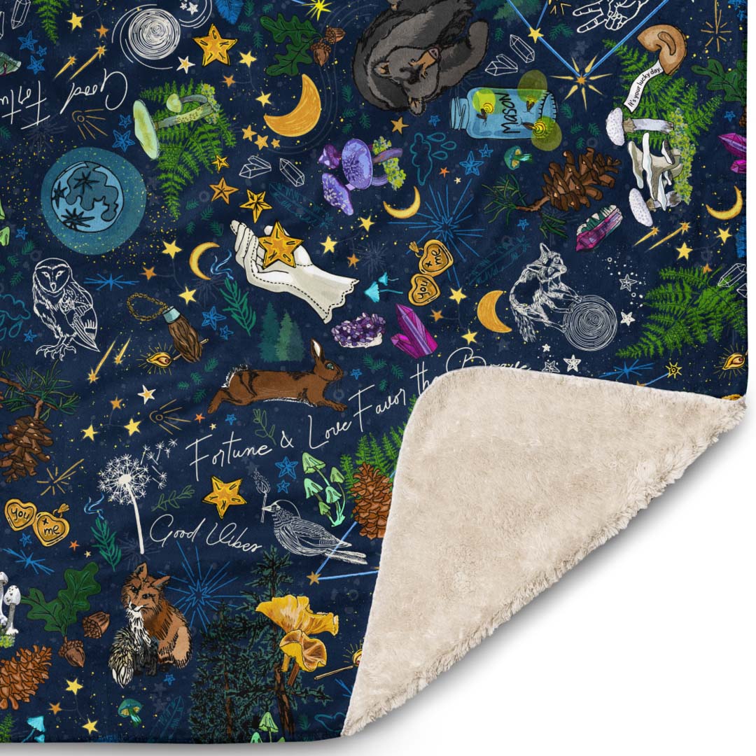 Close-up of the corner of the Under the Stars blanket. The illustrations include lucky constellations, shooting stars, wild mushrooms, ferns, rabbits, bears, foxes, crystals, crescent moons, deer, and other mystical lucky charms.