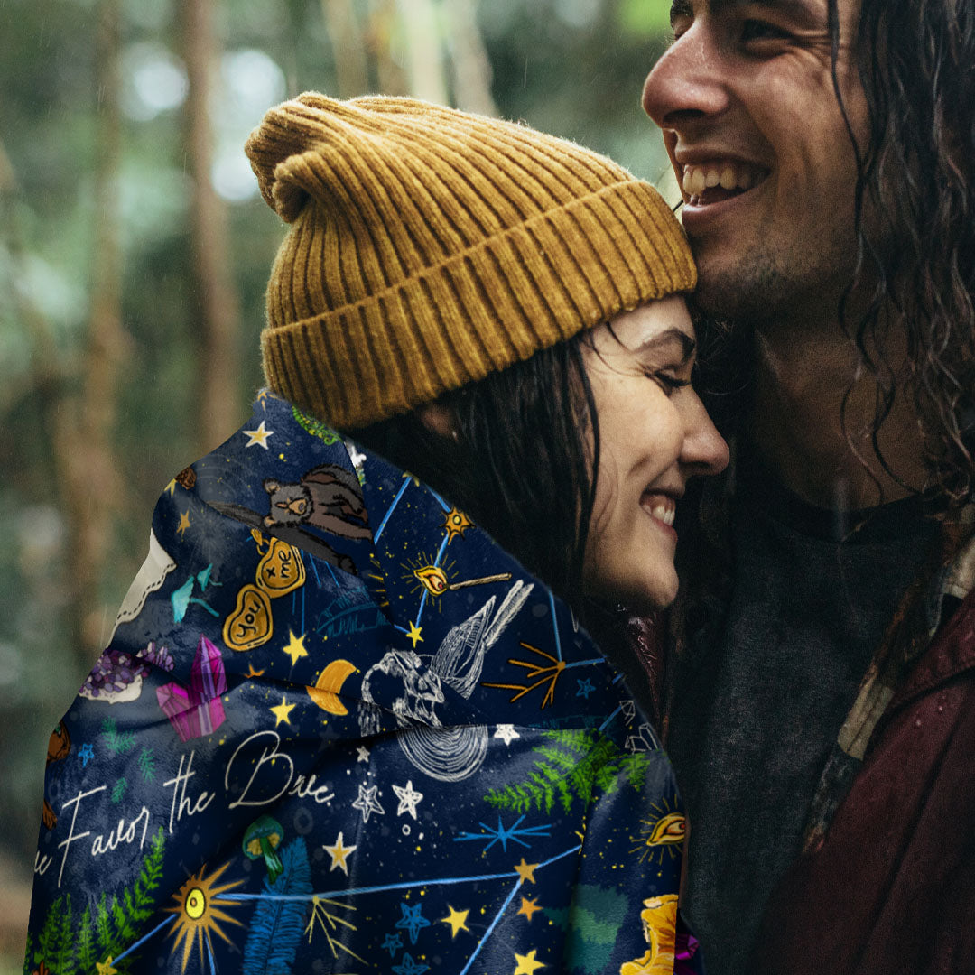 Two people embracing outdoors. The young woman is wearing a mustard yellow beanie, with the Under the Stars sherpa blanket draped around her shoulders