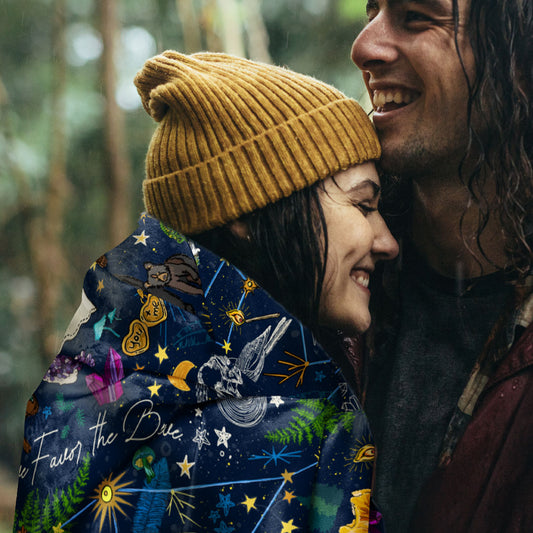 Two people embracing outdoors. The young woman is wearing a mustard yellow beanie, with the Under the Stars sherpa blanket draped around her shoulders