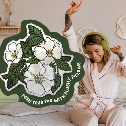 This image shows a photo of the rose-shaped pillow in a green organic shape. Inside the shape are the words "Posh your pad with plushy pillows". In the background, there is a photo of a woman in pajamas wearing headphones and sitting on a bed.