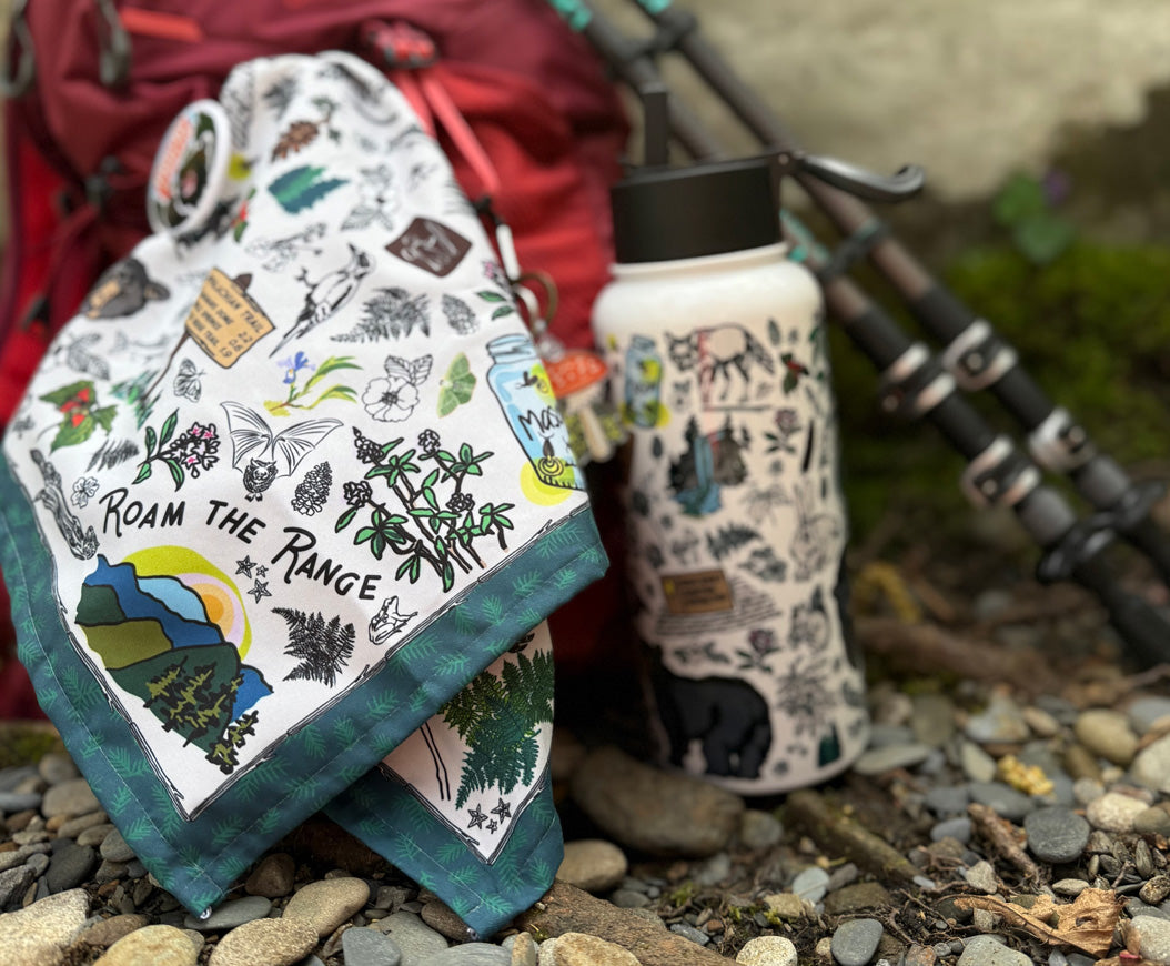 Photo of two Smoky Mountains collection products. It includes the bandana and insulated water bottle. They are sitting on rocks beside hiking poles and reck backpack.